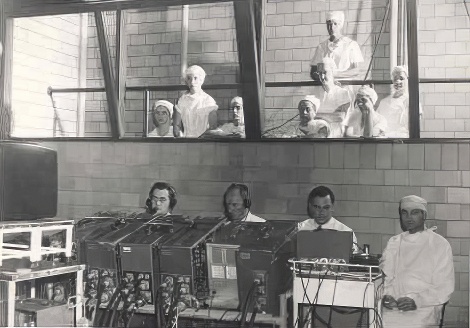 Four men sit in front of TV equipment. Several people watch them from a viewing area above.