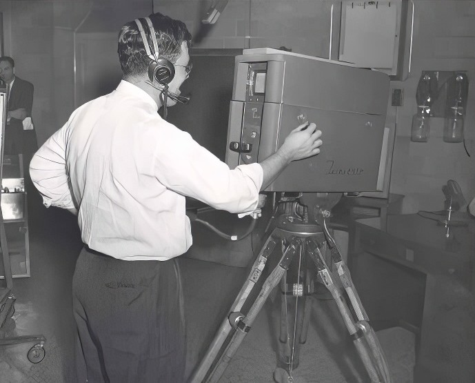 A man wearing a headset stands behind a large camera.