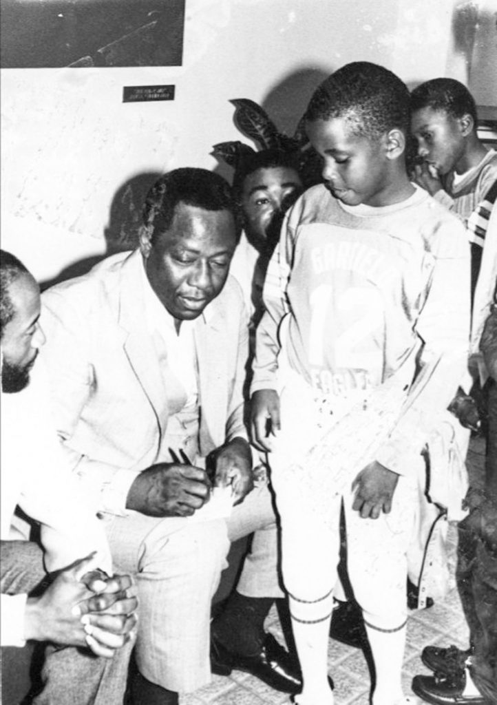 Hank Aaron signs a piece of paper while a young boy in a baseball uniform waits.