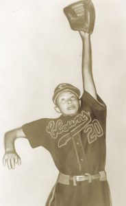 A woman is mid-jump. She is wearing a Clowns baseball uniform with the number 20 on it and has a baseball mitt on her outstretched hand.