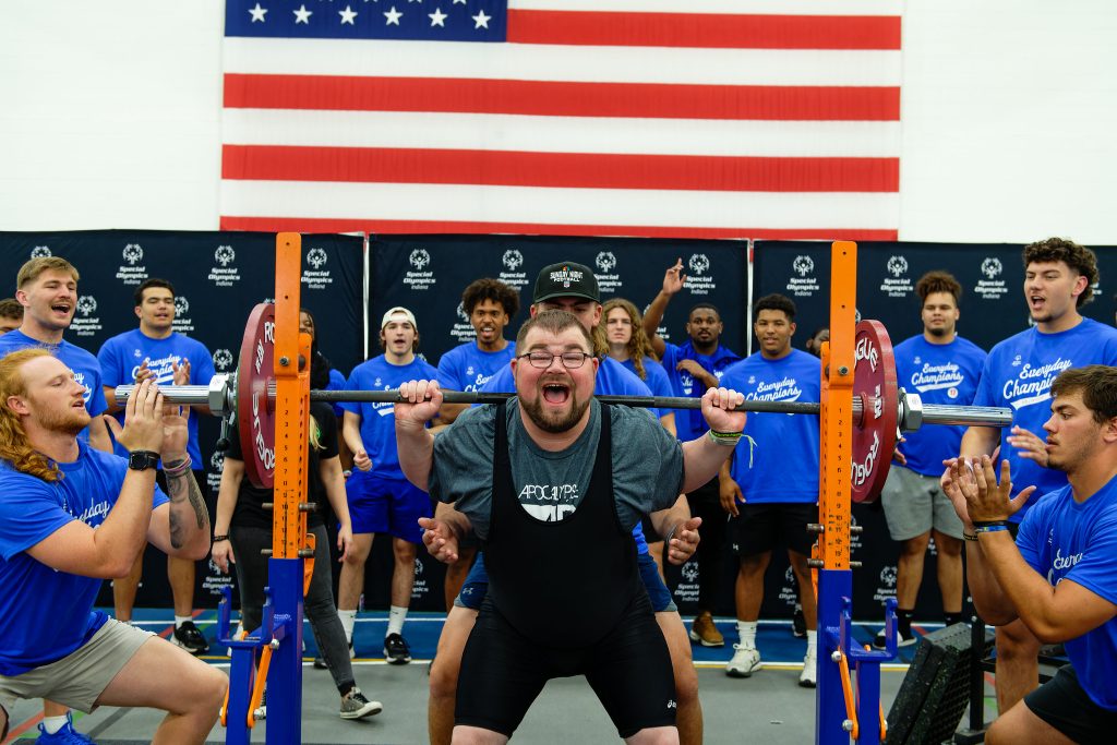 A young man performs a powerlifting squat while a group of individuals cheer him on from behind.