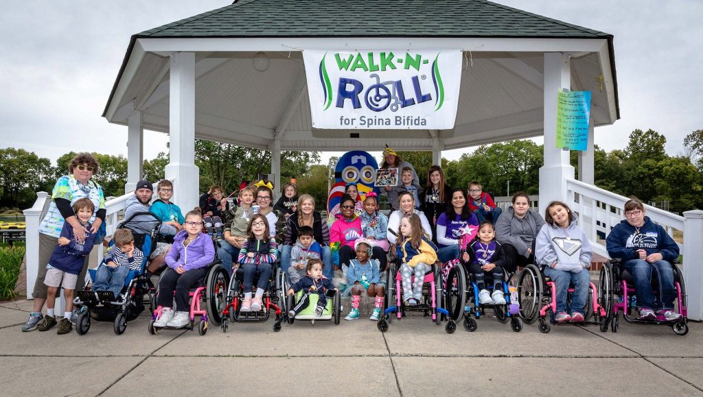 Group photo of children and adults gathered under a pavilion with a banner reading “Walk-N-Roll for Spina Bifida,” many using wheelchairs and mobility devices.