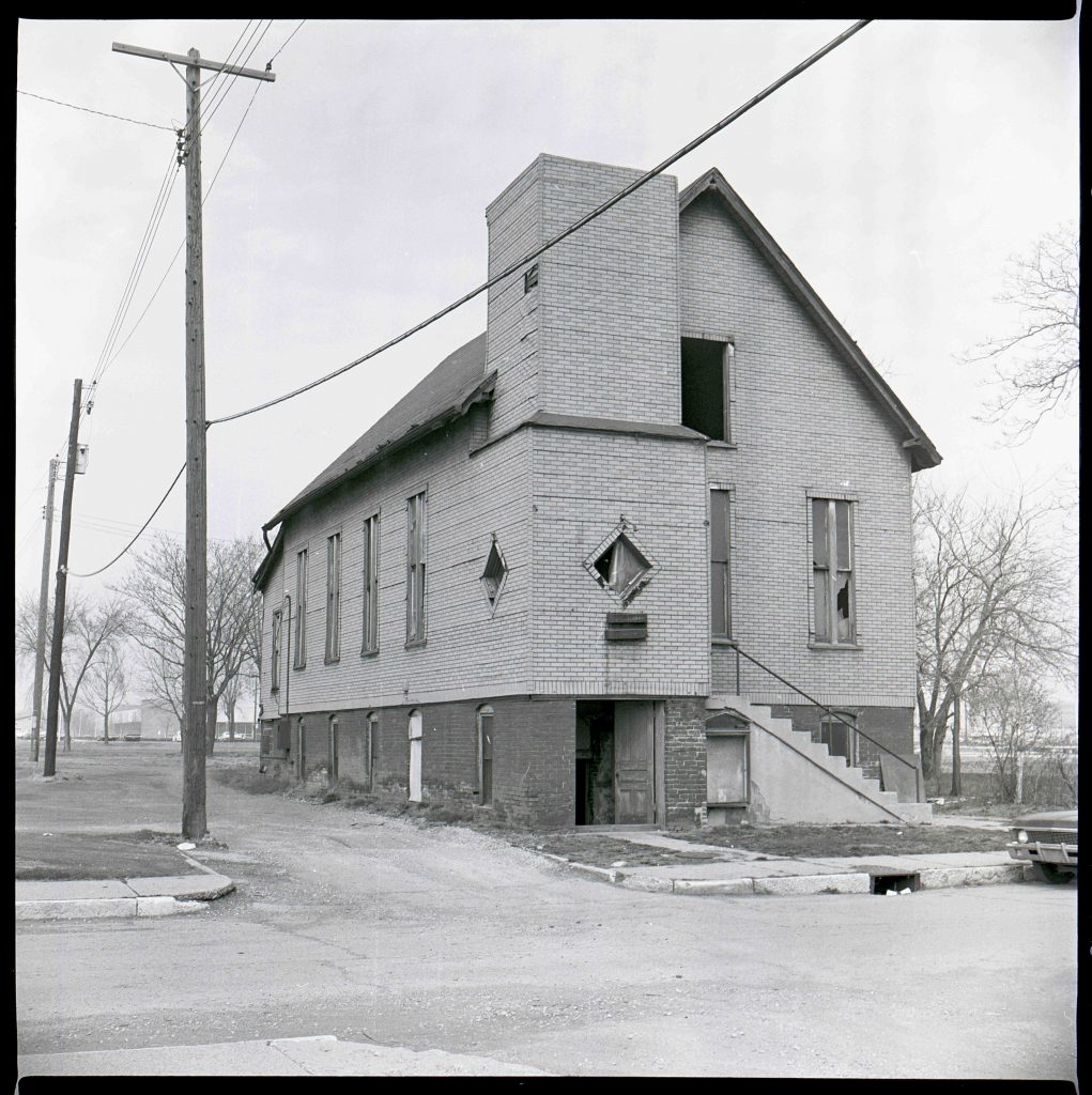 Black-and-white photo of a two-story brick church with boarded and broken windows on a corner lot, with utility poles, bare trees, and a worn street in the foreground.