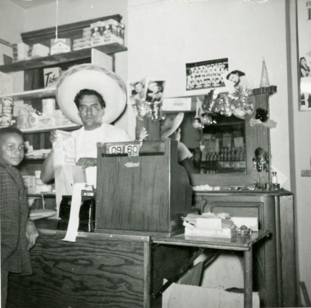 A man stands behind the register at a market. He is placing a carton of milk in a paper bag. A young person waits at the counter for his purchase.