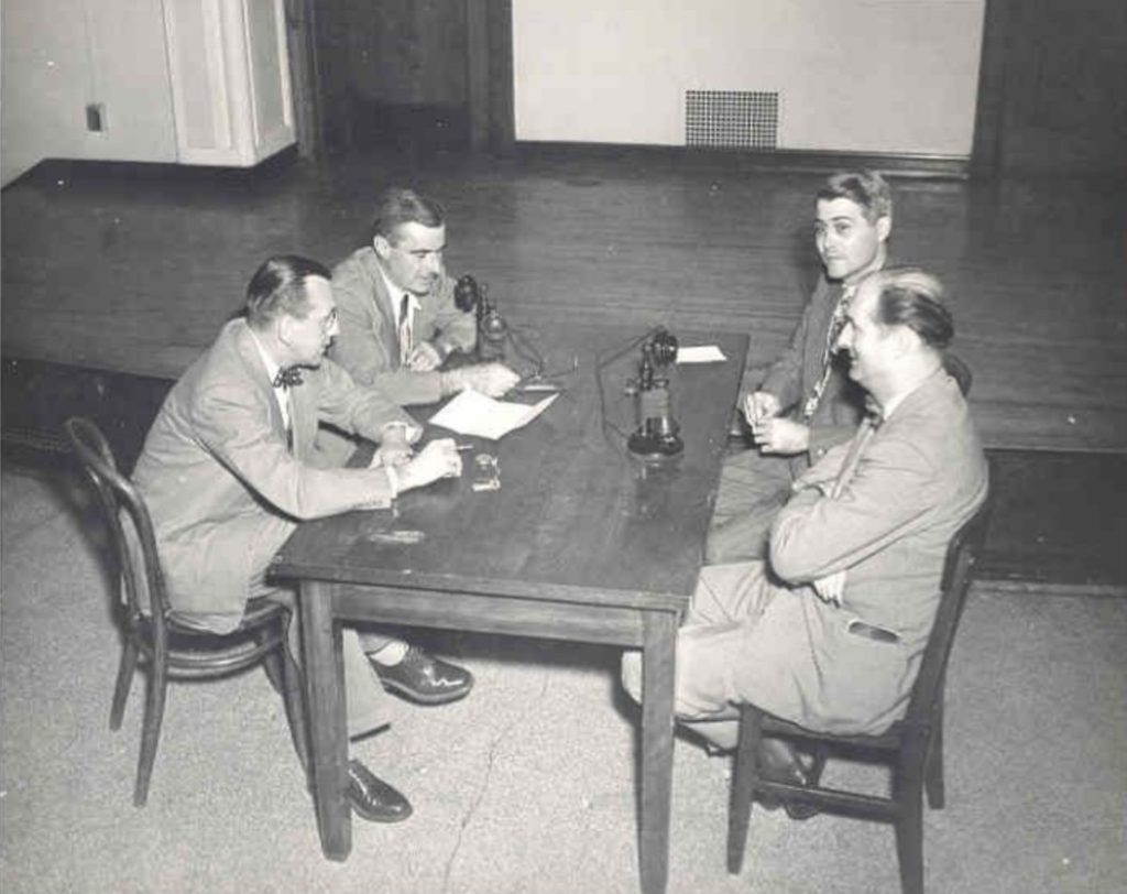 Four men sit around a table. There are two telephones on the table.