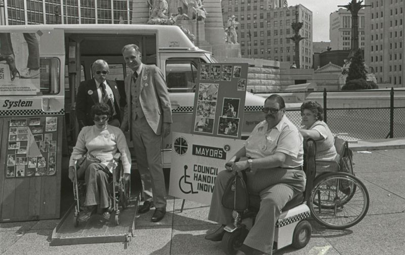 A group of wheelchair users and Mayor Hudnut stand together in front of an accessible van. There are several poster boards set up around them, including one that names them as the "Mayor's Council of Handicapped Individuals."