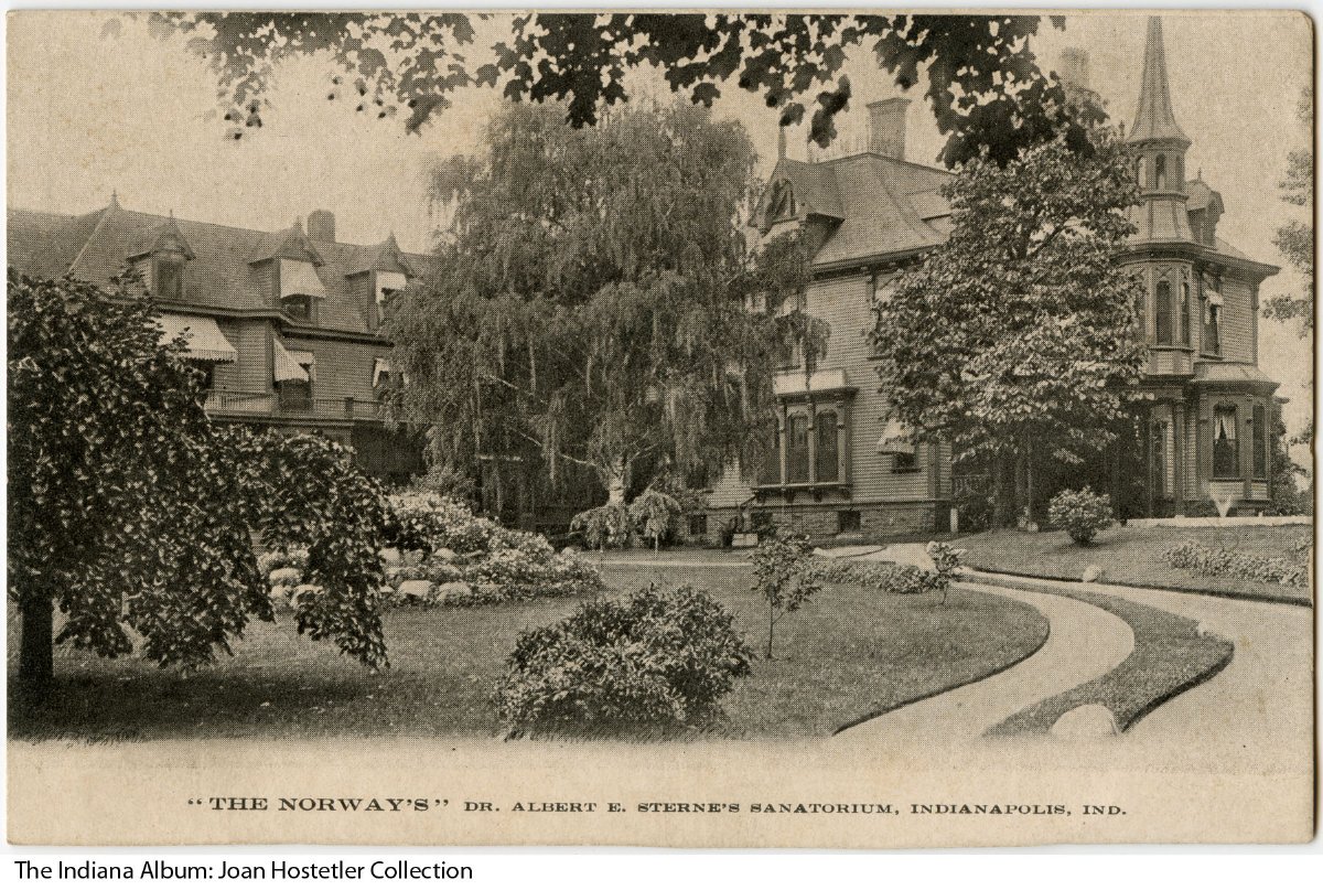 A sprawling brick building with a turret near the entrance and a paved path leading up to the house. Several trees and shrubs dot the front lawn.