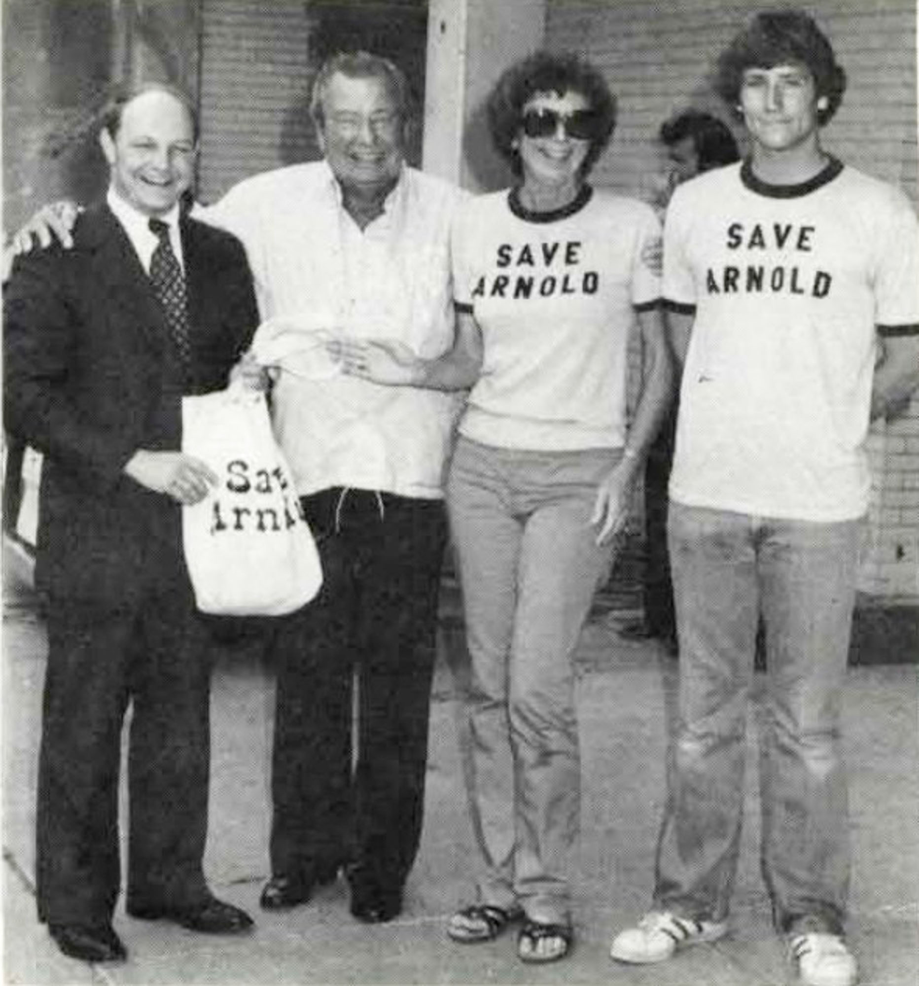 Four people stand together. Two people are wearing shirts that say "Save Arnold" and one person is holding a tote bag with the same phrase.