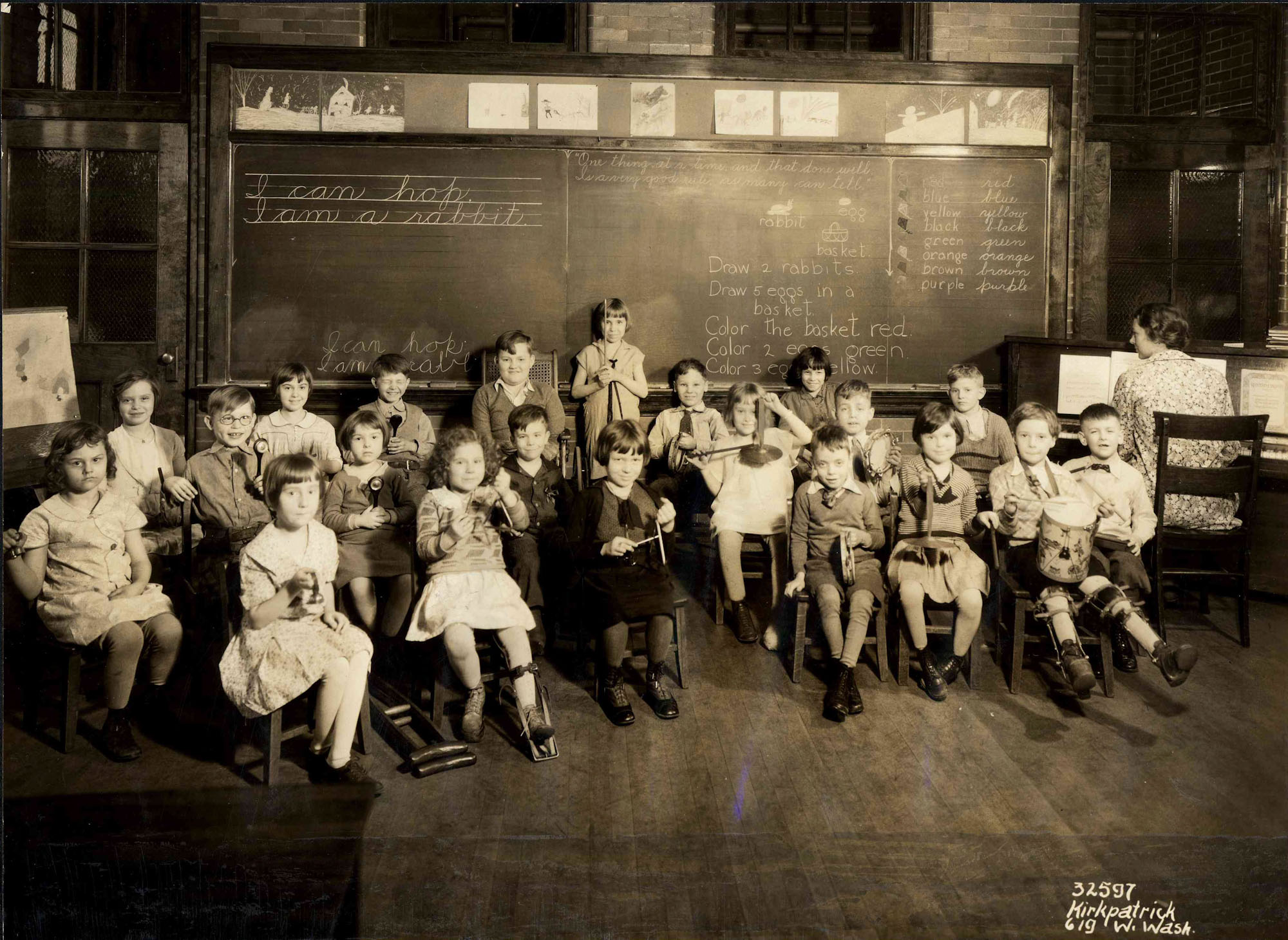 A group of young students sit in a classroom. They all hold instruments, and an adult is seated at a piano. Some of the children wear leg braces. One child has crutches on the floor next to her, and another child sits in a wheelchair.