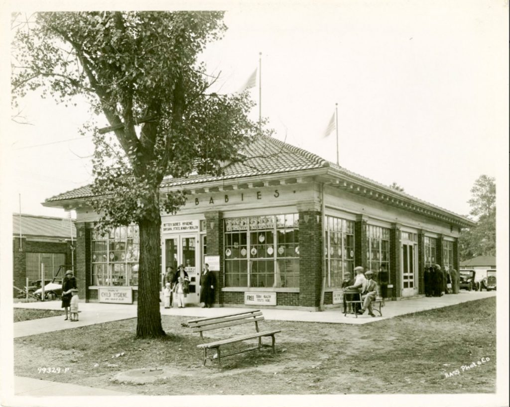 Signs around the building credit the State Board of Health as the sponsor for the exhibition. Pictures of children are hung in the windows. Two little boys in sailor suits are standing at the entrance. A group of adults are looking in the back windows.