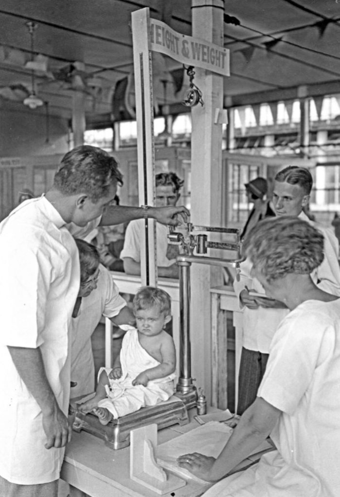 A small child sits on a scale as a man in a lab coat checks her weight. Several other staff aid the man in keeping the child still. One has a notepad in his hand.
