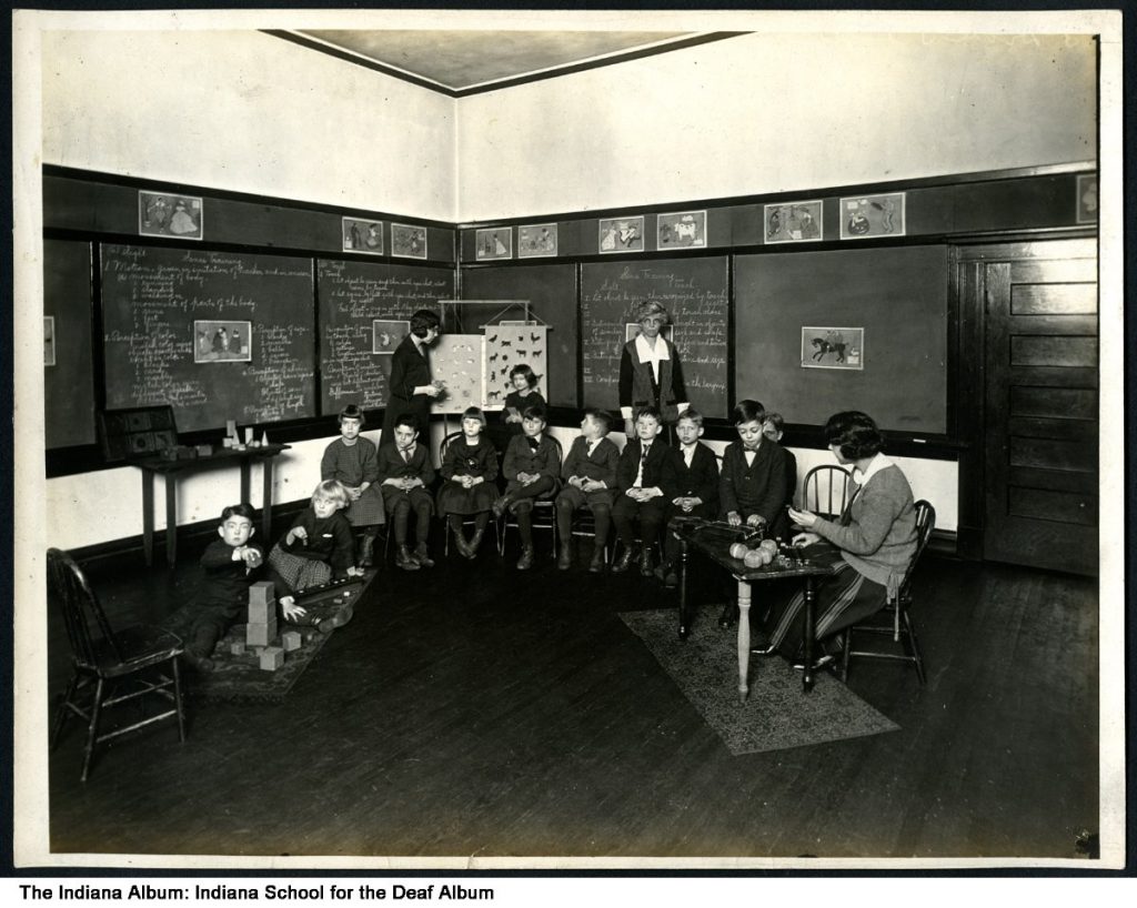 A group of young students sit in a semi-circle in a classroom. There are three teachers working with the students.