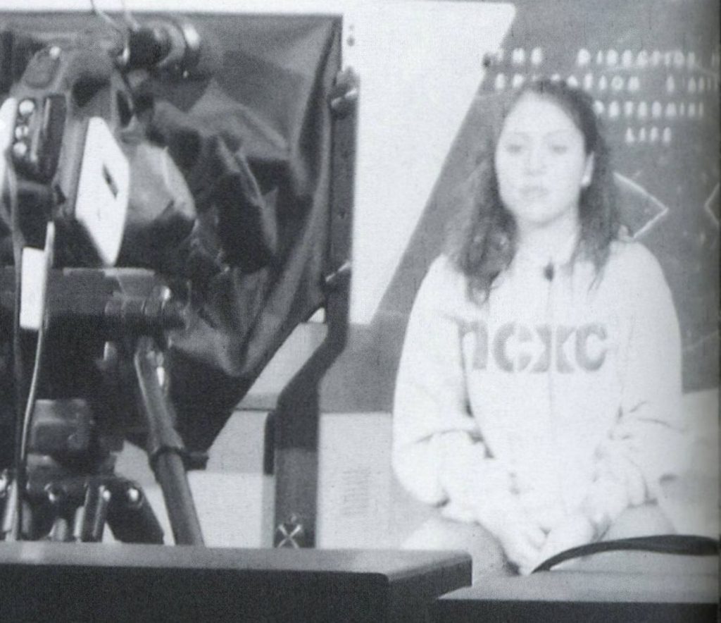 A student sits on a tv set. A camera is pointed at her.