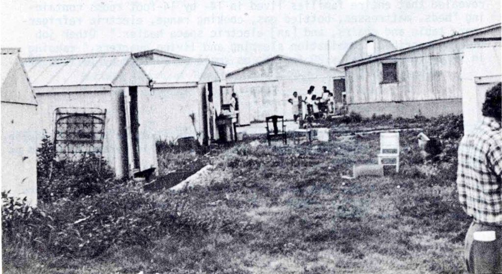 Black-and-white photograph of a migrant camp, showing rows of small shed-like structures and larger buildings separated by grassy, unpaved ground. Several people stand near a building in the background, and a partially visible person appears at the right edge of the image.