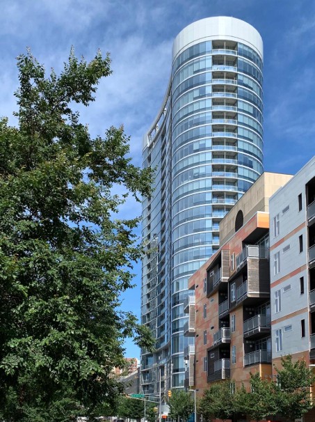 Color photograph of a modern high-rise residential tower with a curved glass façade and rounded top. A shorter apartment building and trees line the street in the foreground beneath a blue sky.