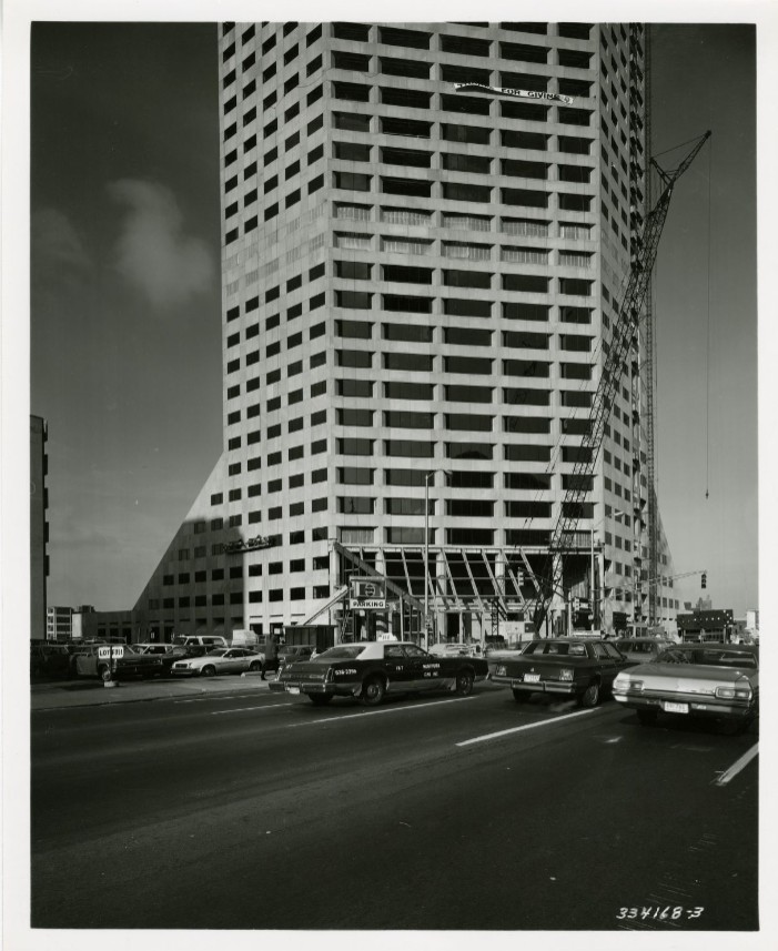 Black-and-white photograph of a tall high-rise office tower under construction beside a city street. A large crane stands along the building's facade, while several cars drive past in the foreground. The lower floors feature an angular, sloping section and an open entrance area.