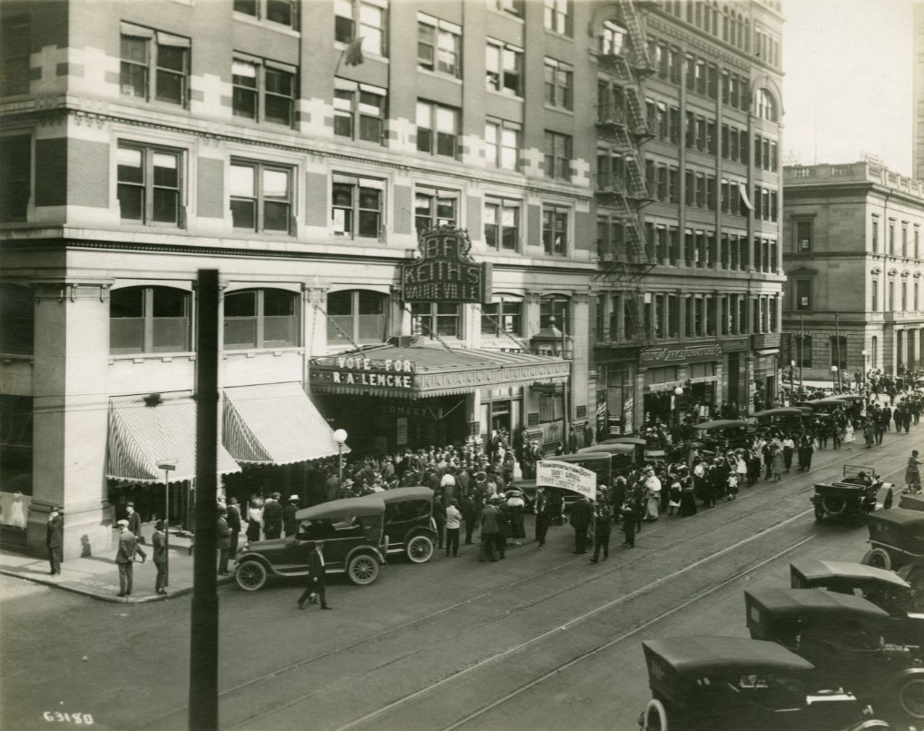 Historic street scene outside B.F. Keith’s Theater in downtown Indianapolis. A crowd gathers beneath the theater marquee while early automobiles line the curb along a street with streetcar tracks. Multi-story commercial buildings with large windows and fire escapes surround the theater. Visible signs include “B.F. Keith’s Vaudeville” and a political banner reading “Vote for R. A. Lencke.”