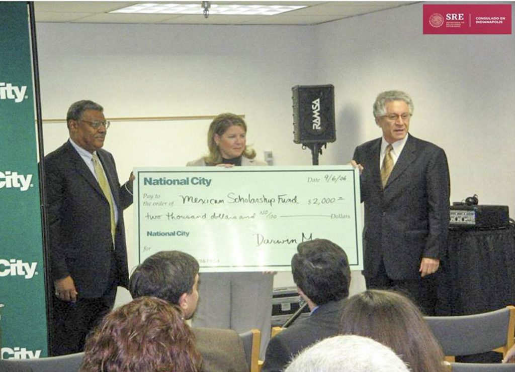 People stand before an audience while presenting an oversized $2,000 check from National City to the Mexican Scholarship Fund at a community event. A banner and speaker are visible in the background.