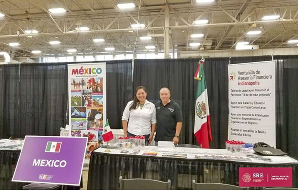 Information booth at the Indiana Latino Expo featuring Mexico-themed displays, Mexican flags, and educational materials. Two people stand behind a table between banners promoting Mexican community services and financial education programs.