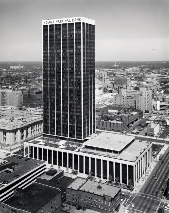 Aerial view of the Indiana National Bank Tower rising above downtown Indianapolis, with surrounding office buildings, streets, and parking areas visible below.