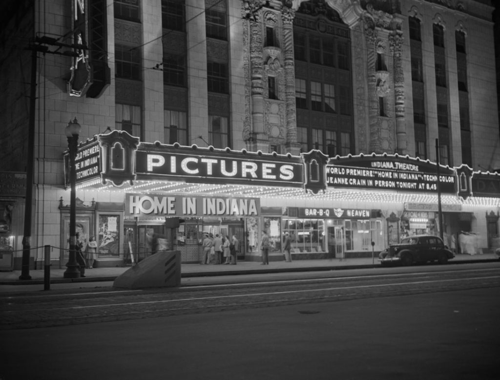Historic nighttime view of the Indiana Theatre in downtown Indianapolis. The theater’s illuminated marquee advertises Home in Indiana and a world premiere event, while people gather near the entrance. Streetcar tracks run along the foreground, and an automobile is parked at the curb beneath the ornate theater façade lined with glowing lights.