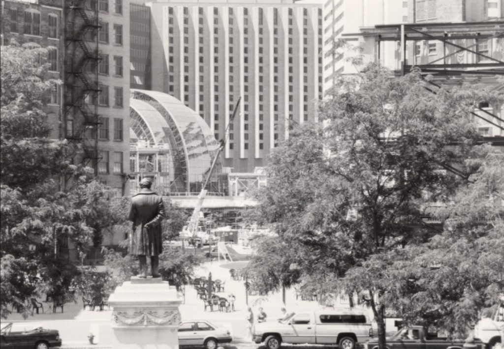 Black-and-white photograph of downtown Indianapolis showing the Soldiers and Sailors Monument statue in the foreground, with trees, parked vehicles, and construction activity around the Artsgarden visible between surrounding high-rise buildings.
