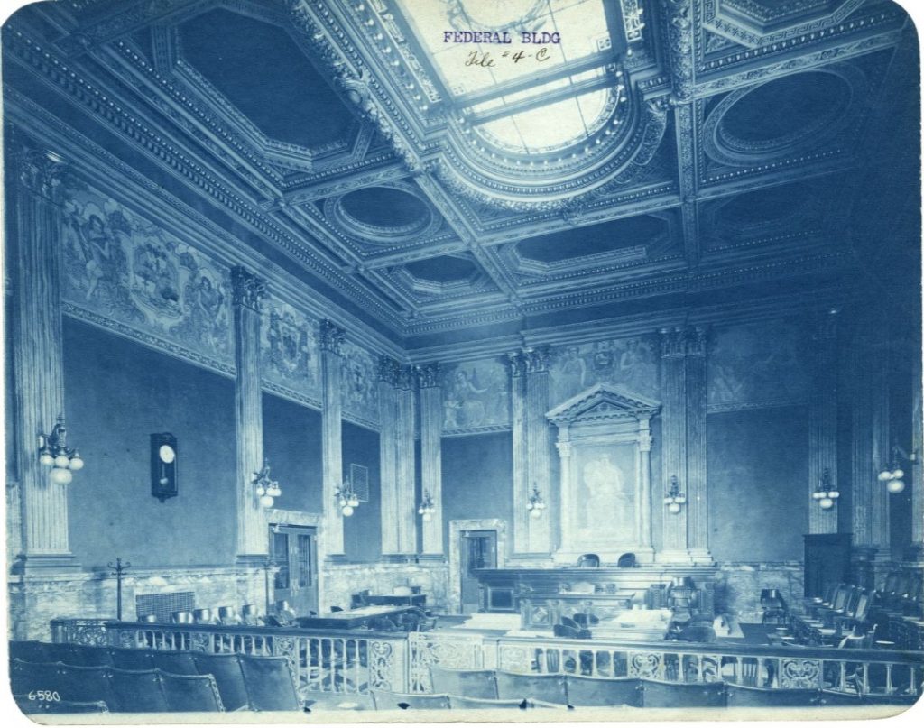 Historic courtroom interior with an ornate coffered ceiling, decorative murals, marble columns, and a raised judge’s bench. Rows of seating and carved railings fill the grand chamber.