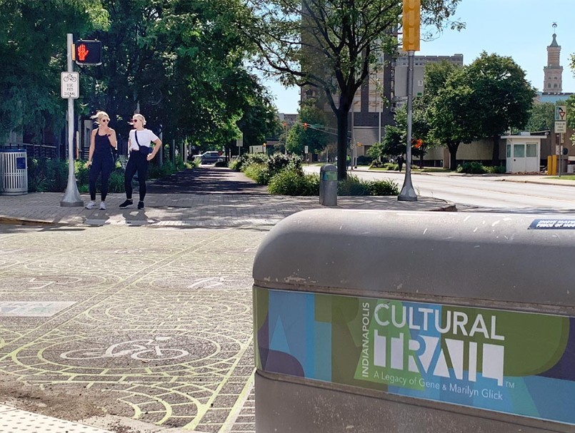 Color photograph of the Indianapolis Cultural Trail showing a patterned bike and pedestrian path at a downtown intersection. Two pedestrians stand near a crosswalk signal, while trees line the trail and the foreground includes a trail sign with the text “Indianapolis Cultural Trail.”