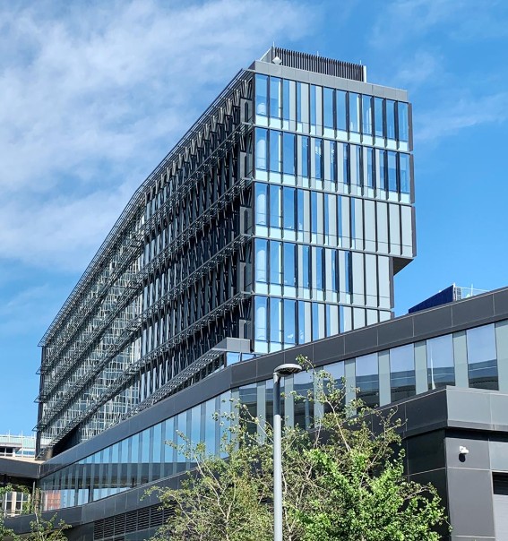 Color photograph of a modern glass-and-steel office building with reflective windows and an angular design. Trees and a streetlight stand in the foreground beneath a blue sky with scattered clouds.
