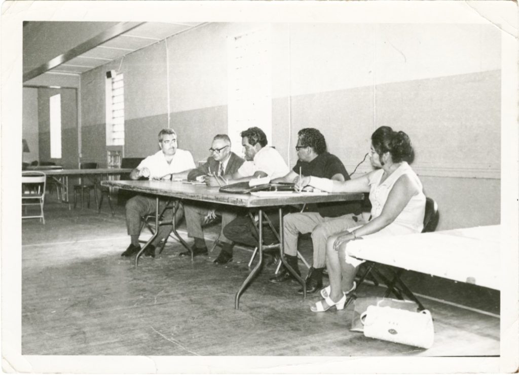 Black-and-white photograph of six people seated around folding tables in a meeting room. Several participants take notes or converse during a discussion, while empty tables and chairs line the background.
