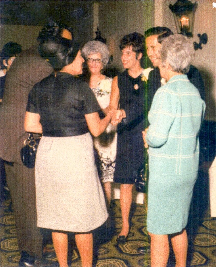 Color photograph of several people gathered at a social event, standing in conversation in an indoor reception space. Attendees wear formal or semi-formal clothing and are grouped in discussion beneath wall-mounted light fixtures.