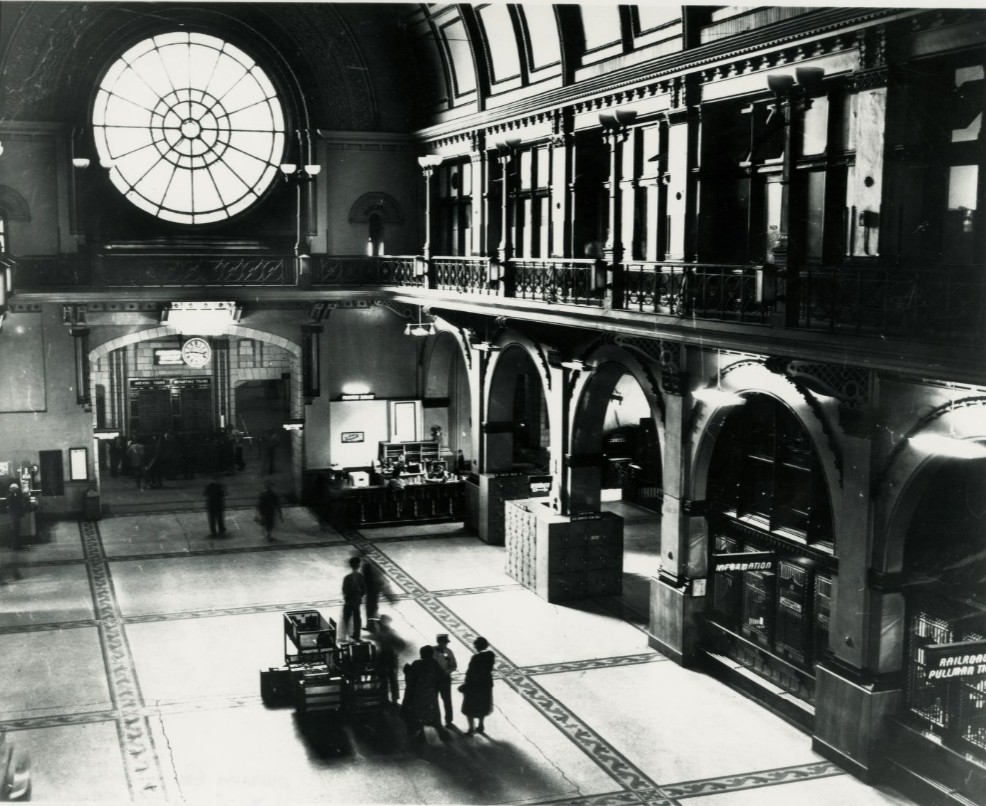Interior view of Union Station’s Great Hall, featuring a large circular window above an arched entrance, ornate balconies and columns, ticket and information counters, and a few travelers standing on the expansive floor below.
