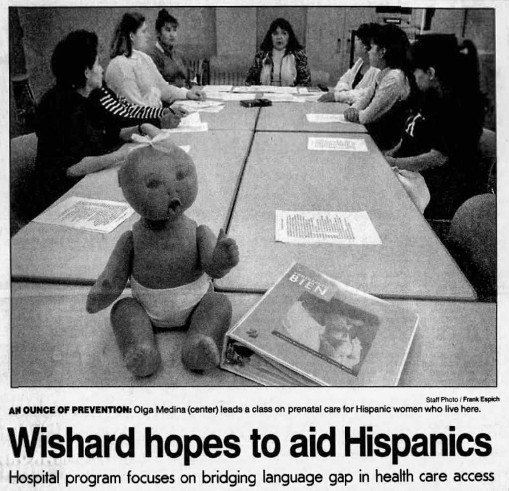 Newspaper photo showing a prenatal care class seated around a conference table. A baby doll and a Spanish-language parenting book sit in the foreground. The article headline reads, “Wishard hopes to aid Hispanics,” about a hospital program addressing language barriers in health care.