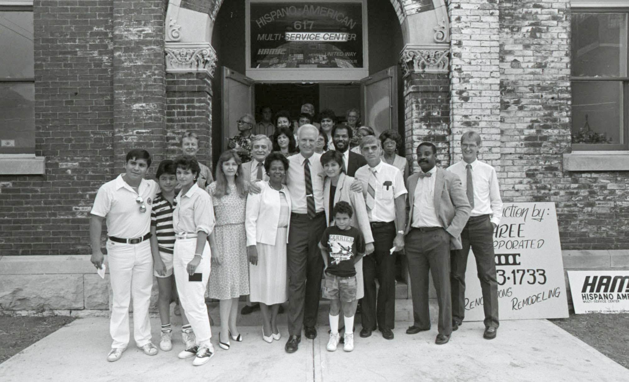 Black-and-white group photograph of adults and children standing on the steps outside the Hispano-American Multi-Service Center. The group poses in front of a brick building with the center’s name displayed above the entrance.