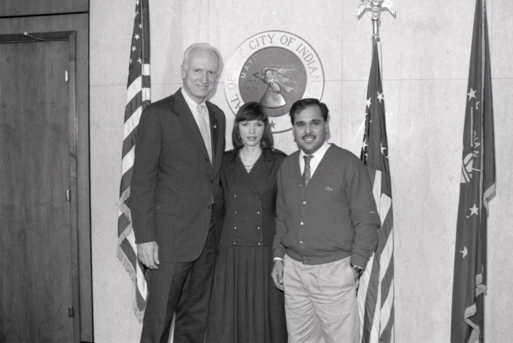 Black-and-white photograph of three people posing in front of the seal of the City of Indianapolis, with U.S. and Indiana flags displayed on either side. The group stands inside a government office or meeting room.