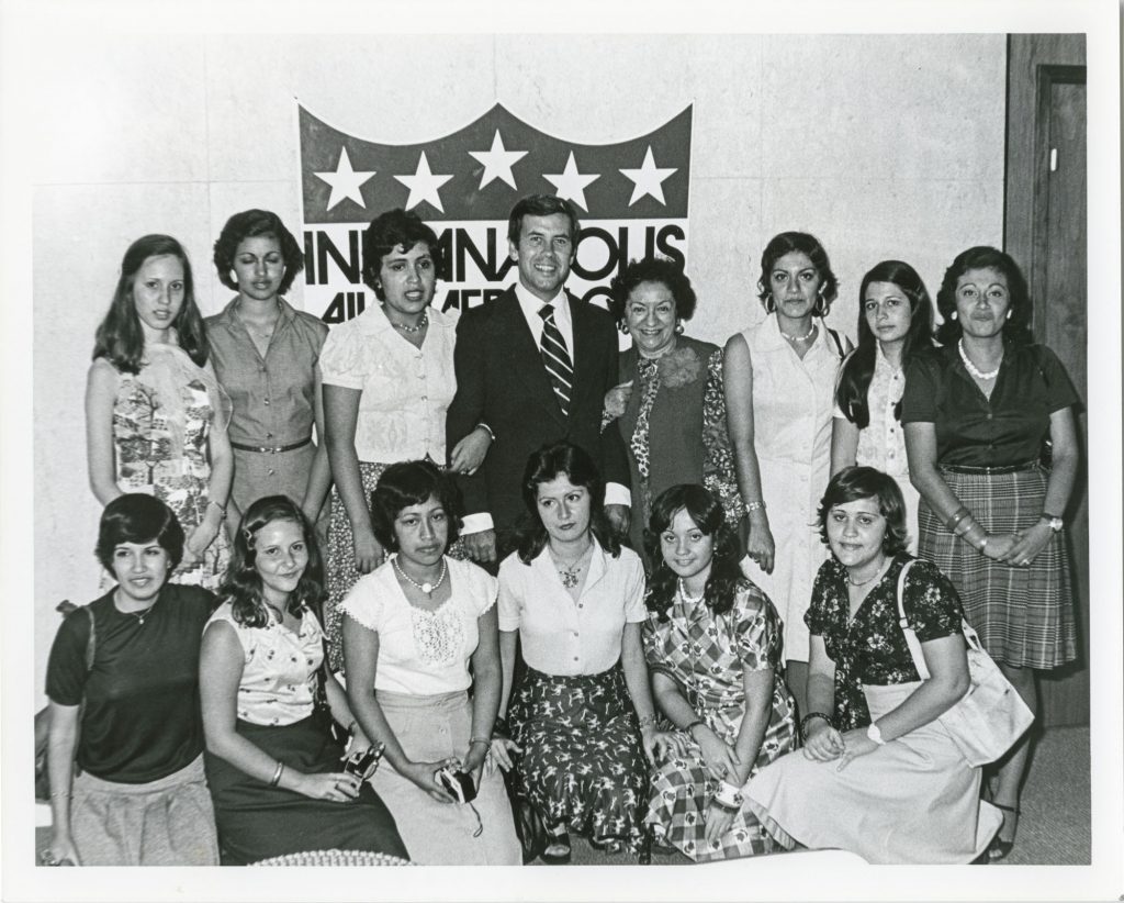 Black-and-white group photograph showing a suited official standing with a group of young people in front of an Indianapolis city government sign. Several people are standing while others are seated, posing for a group portrait inside a public building.