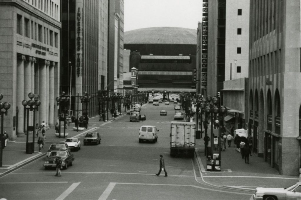 Black-and-white photograph of a downtown street lined with tall office buildings, with cars, a delivery truck, and pedestrians crossing the roadway. Market Square Arena rises in the distance at the end of the street.