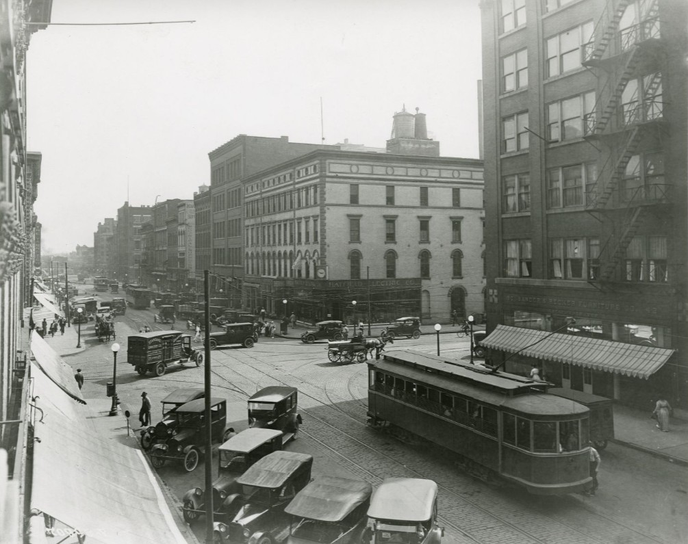 Historic downtown street scene with early automobiles, a streetcar, and pedestrians at a busy intersection lined with multistory commercial buildings. Tracks run through the street, and fire escapes are visible on the building at right.