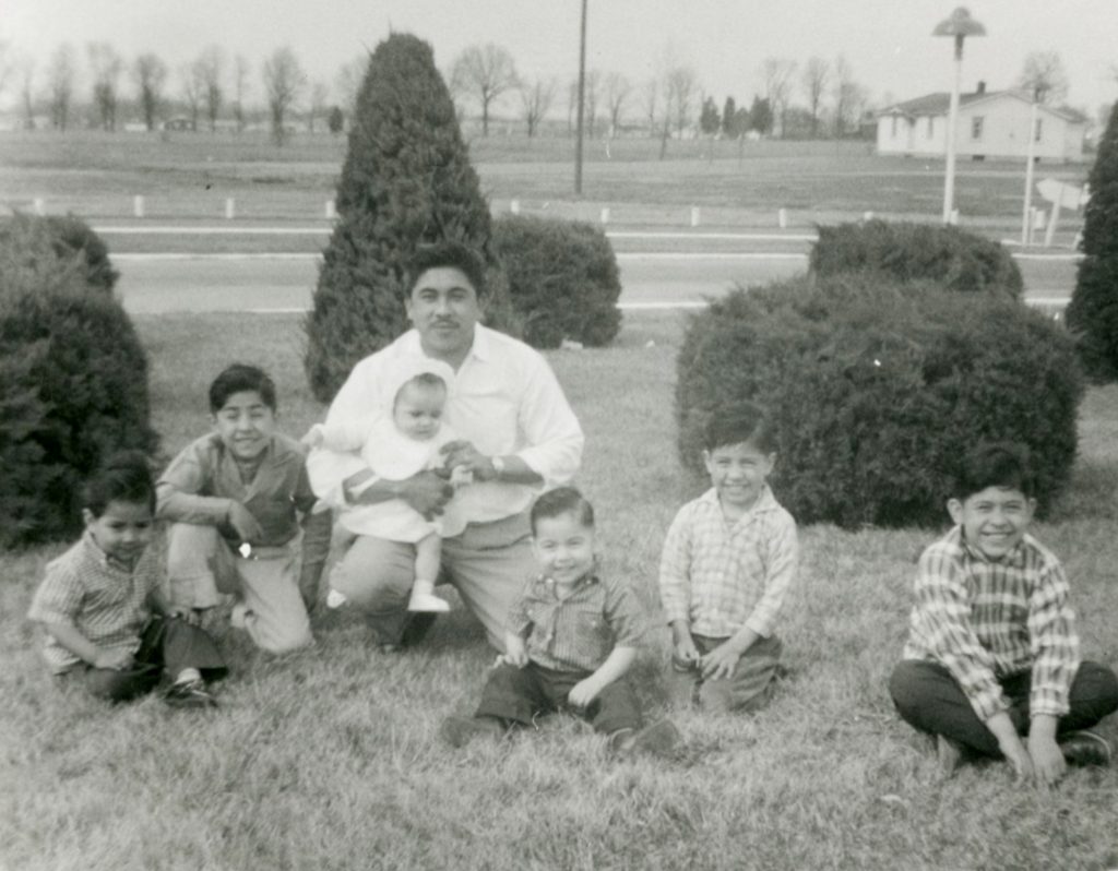 Black-and-white family portrait showing one adult seated on grass holding an infant, surrounded by five young children in a landscaped outdoor area with shrubs, a road, and open fields in the background.