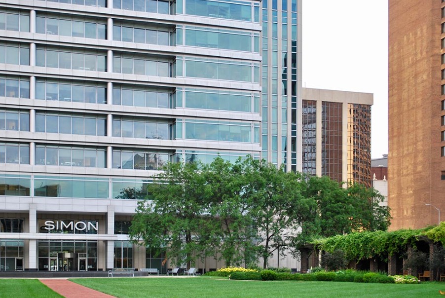 Color photograph of the Simon Property Group headquarters in downtown Indianapolis. The image shows a modern glass office building behind a landscaped lawn with trees, shrubs, benches, and surrounding downtown high-rises.