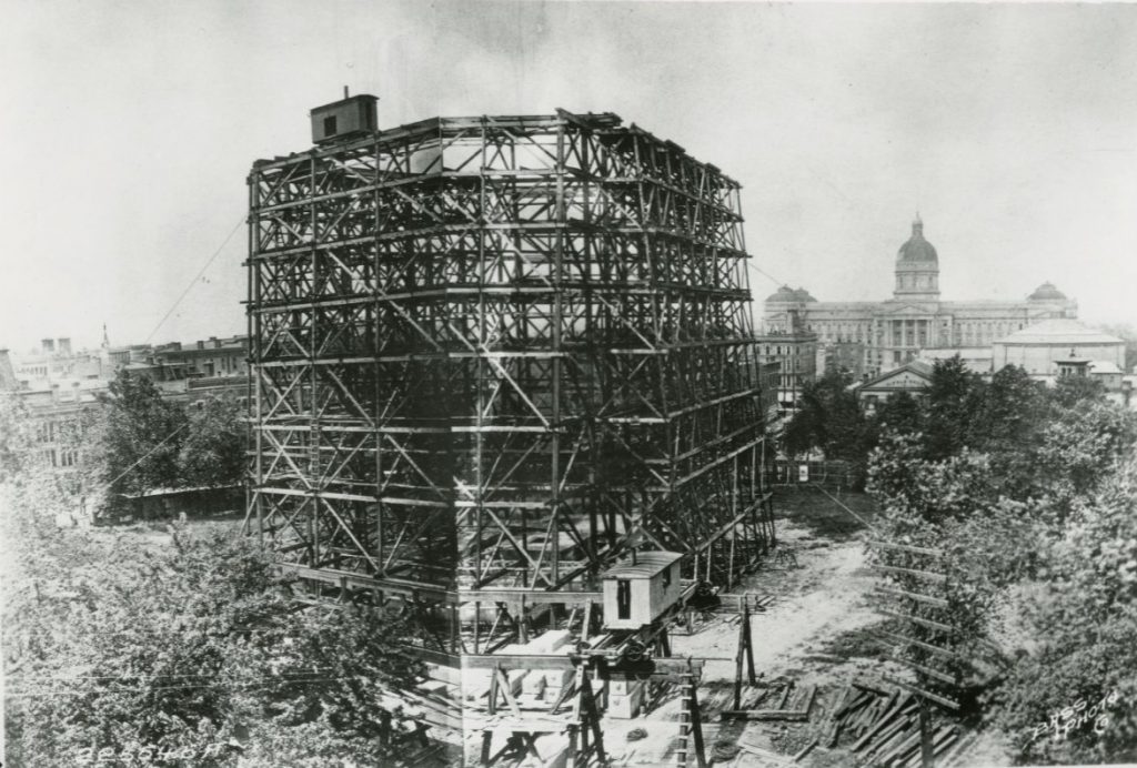 Black-and-white photograph of a large framework under construction in downtown Indianapolis, with the Indiana Statehouse visible in the background. Trees, construction materials, and a small work shed surround the unfinished structure.