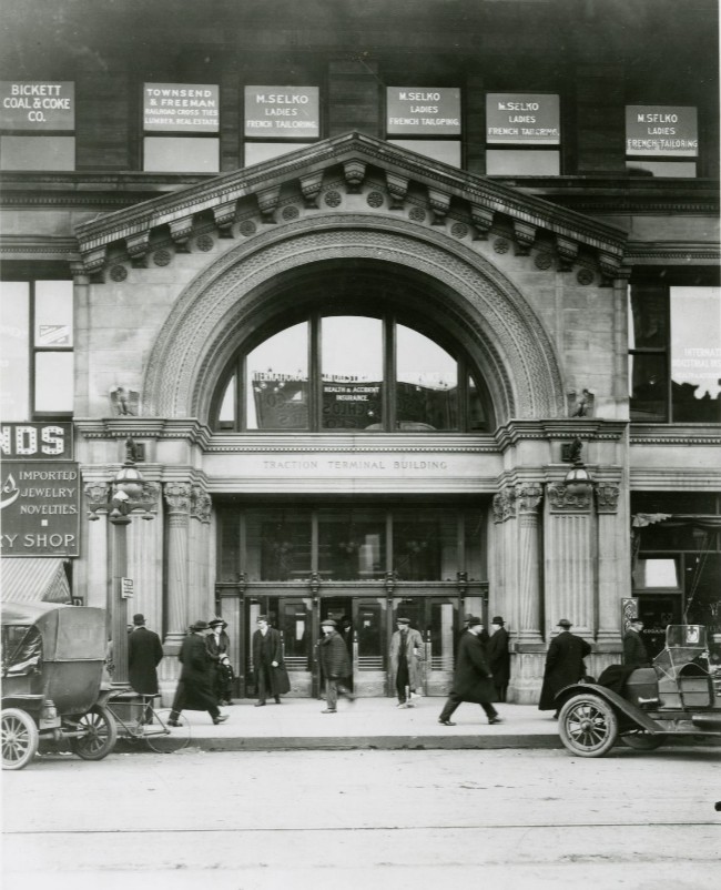 Historic photograph of the Traction Terminal Building in downtown Indianapolis. The ornate stone entrance features a large arched window, with “Traction Terminal Building” carved over the main entrance. Pedestrians walk along the sidewalk in front of the building, while an early automobiles parked at the curb. Business signs are visible in the upper-story windows.