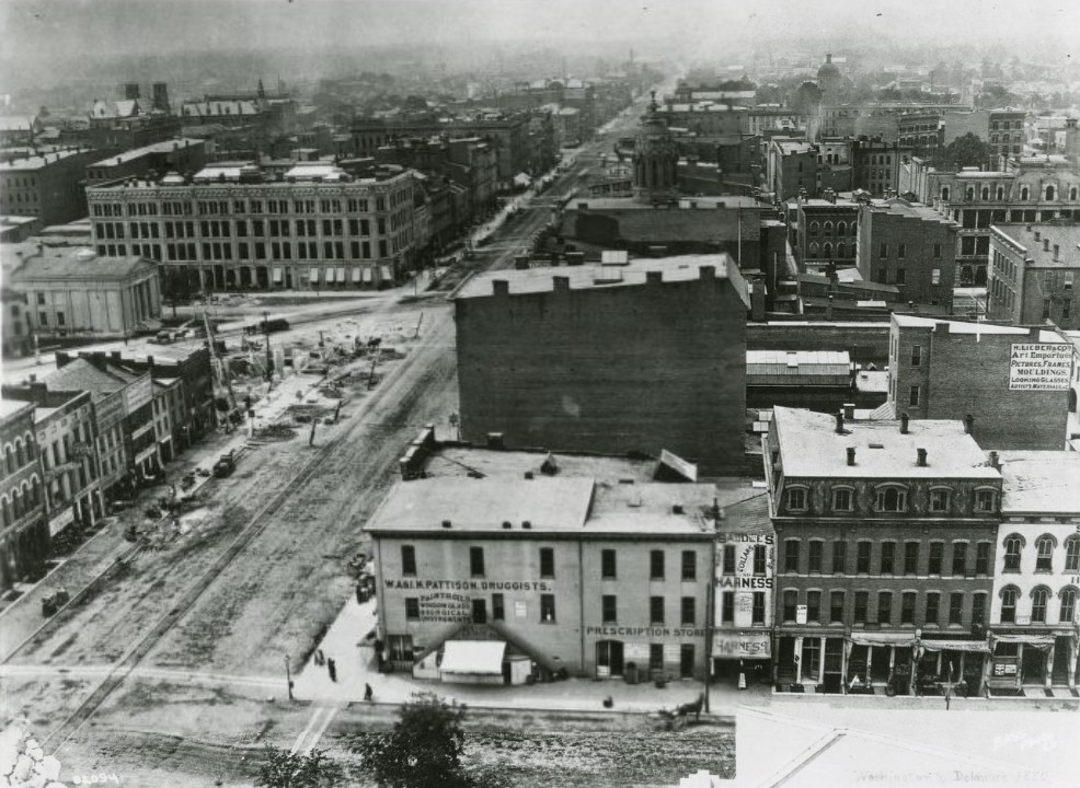 Historic black-and-white aerial view of downtown Indianapolis at Washington and Delaware streets, showing early 20th-century commercial buildings, unpaved streets, streetcar tracks, and a busy urban intersection stretching into the distance.