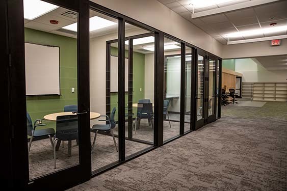 Glass-walled study and tutoring rooms line a carpeted hallway inside the renovated Nora Branch, with tables, chairs, and shelving visible in adjacent library spaces.