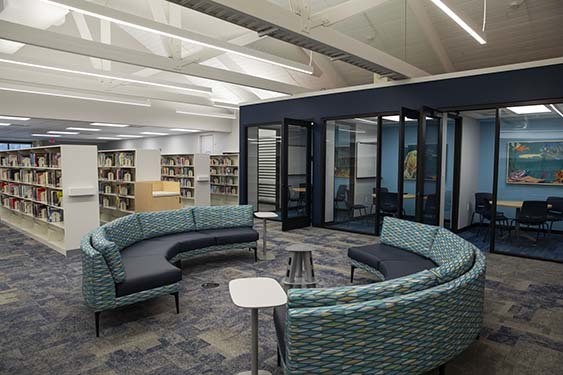 Interior of a library featuring bookshelves, curved upholstered seating, small tables, and glass-walled meeting rooms beneath exposed white ceiling beams.