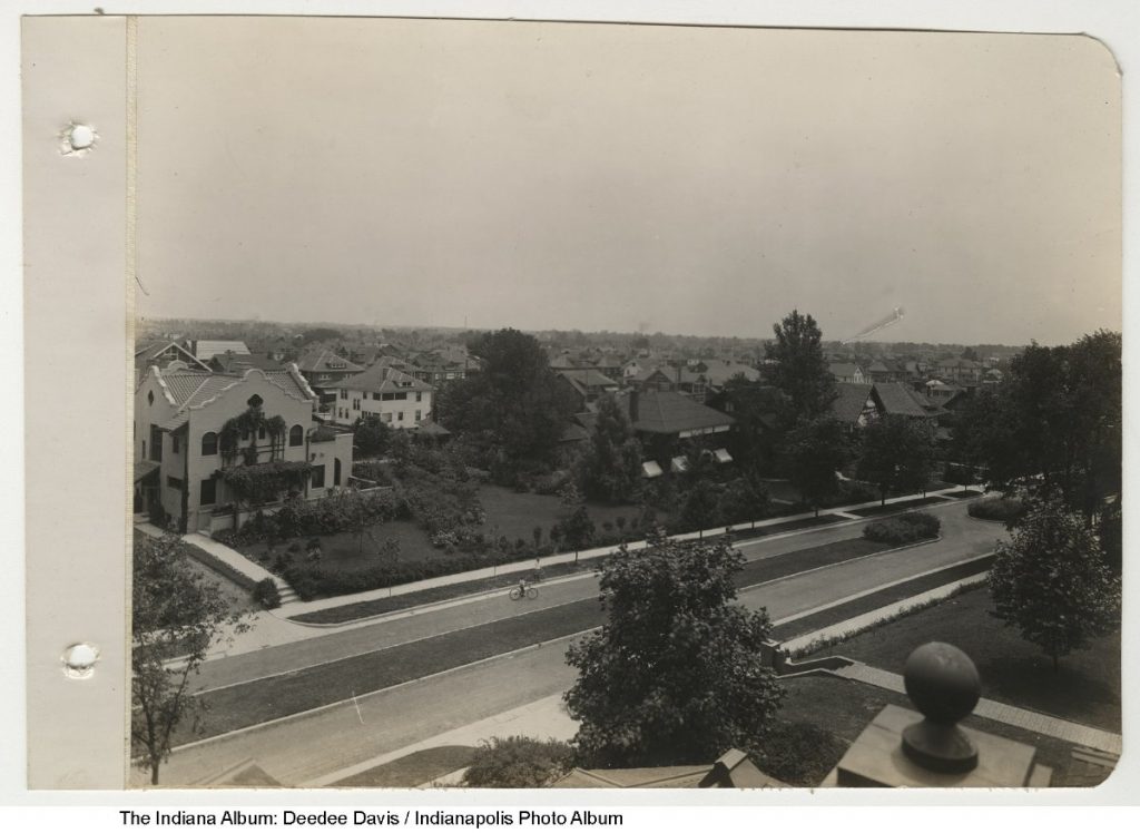 Large stucco house with a decorative curvilinear roofline, arched windows, and climbing vines on the facade, set on a landscaped corner lot along a wide residential street. Black-and-white photograph with surrounding neighborhood homes in the background.