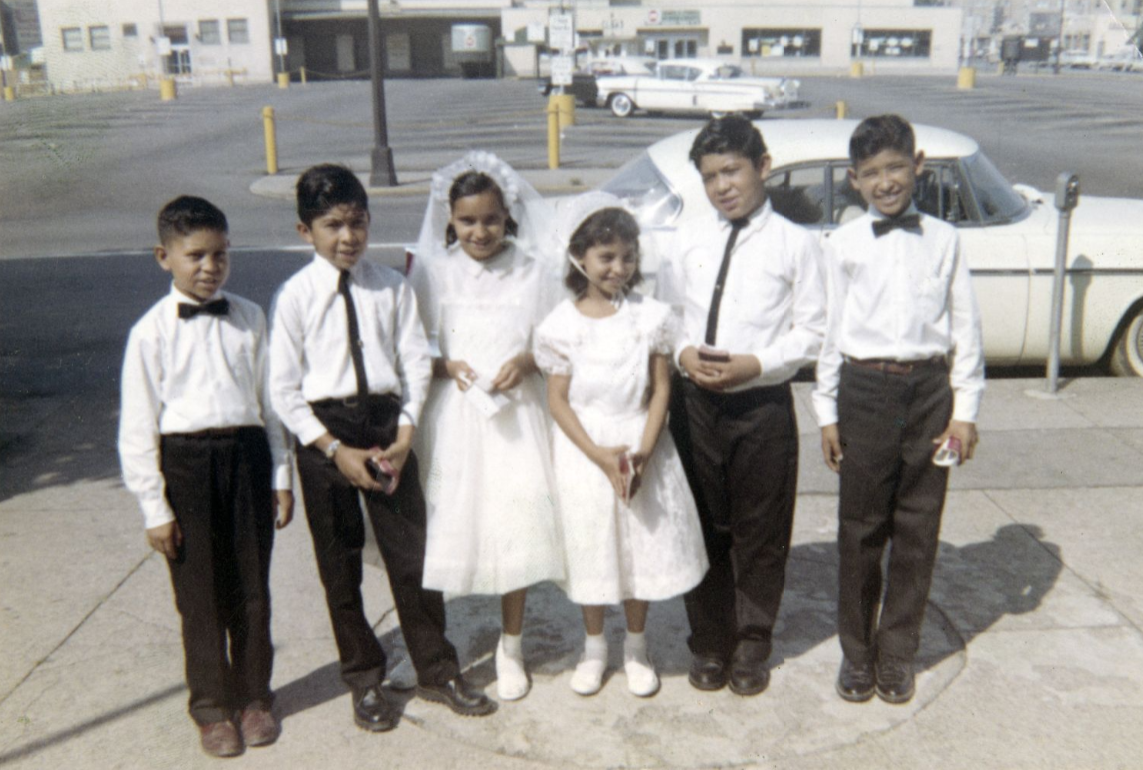 Six children dressed in formal clothing stand on a sidewalk beside a parked car. Two children wear white ceremonial dresses, including one with a veil, while the others wear white shirts, dark trousers, and bow ties or neckties.