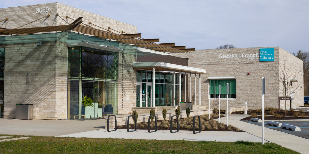 Exterior view of the Glendale Branch of The Indianapolis Public Library, featuring a light stone facade, large glass entrance, covered walkway, bike racks, and landscaping in front of the building.