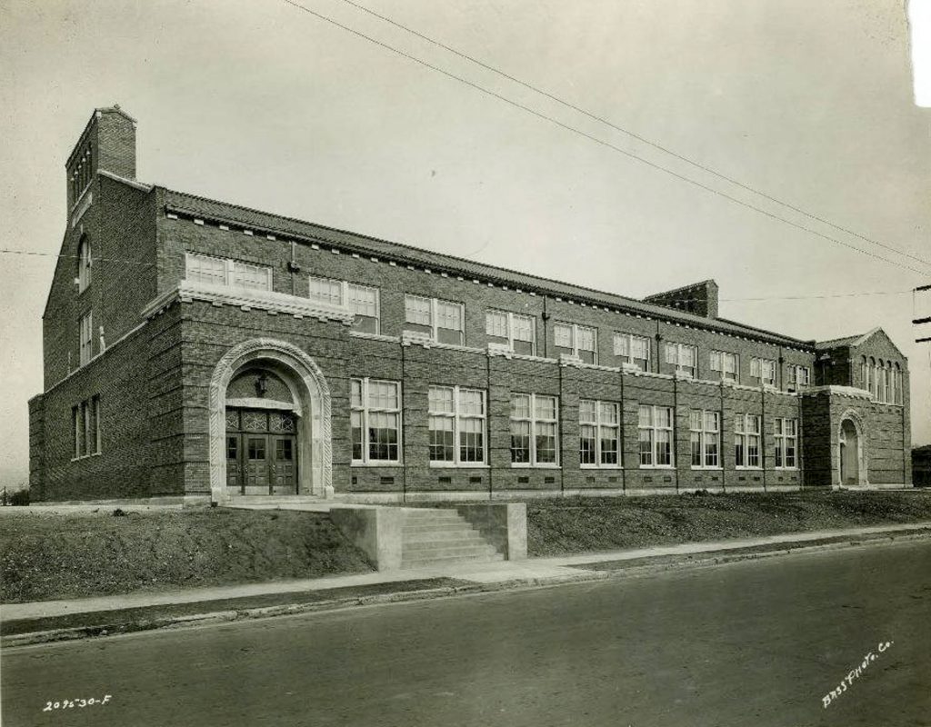 Historic two-story brick school building with arched entrances, large rows of windows, and a broad sidewalk along the street in front.