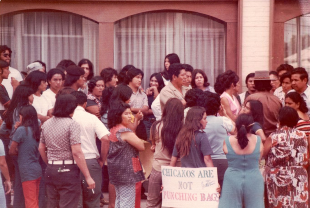 Crowd gathered outside a building during a public demonstration. A protest sign in the foreground reads, “CHICANOS ARE NOT PUNCHING BAGS.”